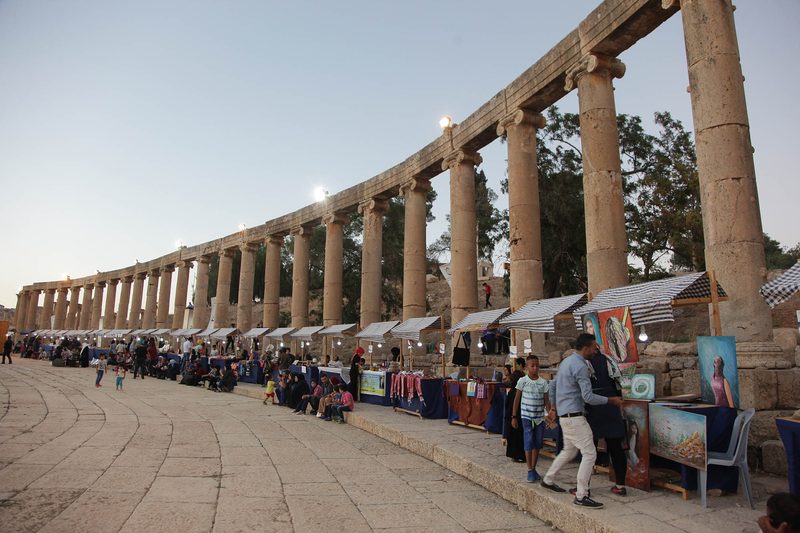 The curved colonnade of the Oval Plaza in Jerash at dusk, with visitors walking among the stalls