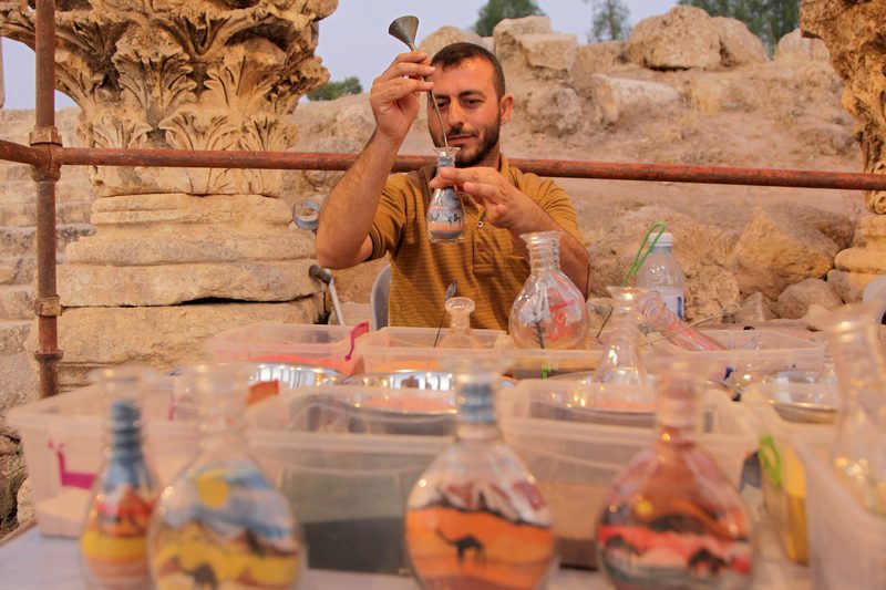 An artist pouring colored sand into a glass bottle beside the ancient columns of Jerash