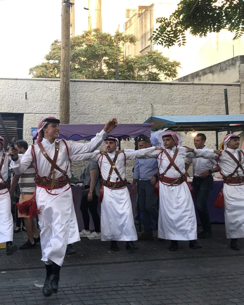 A line of men in white thobes and red shemaghs dancing dabke in a street in Jordan