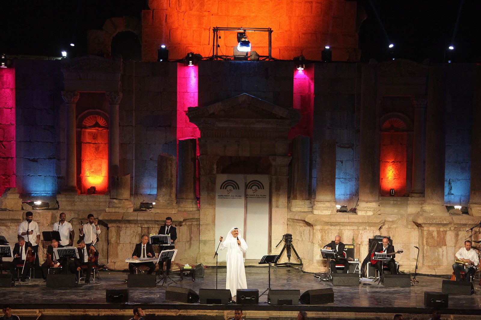 Musicians performing on a lit stage before ancient stone ruins